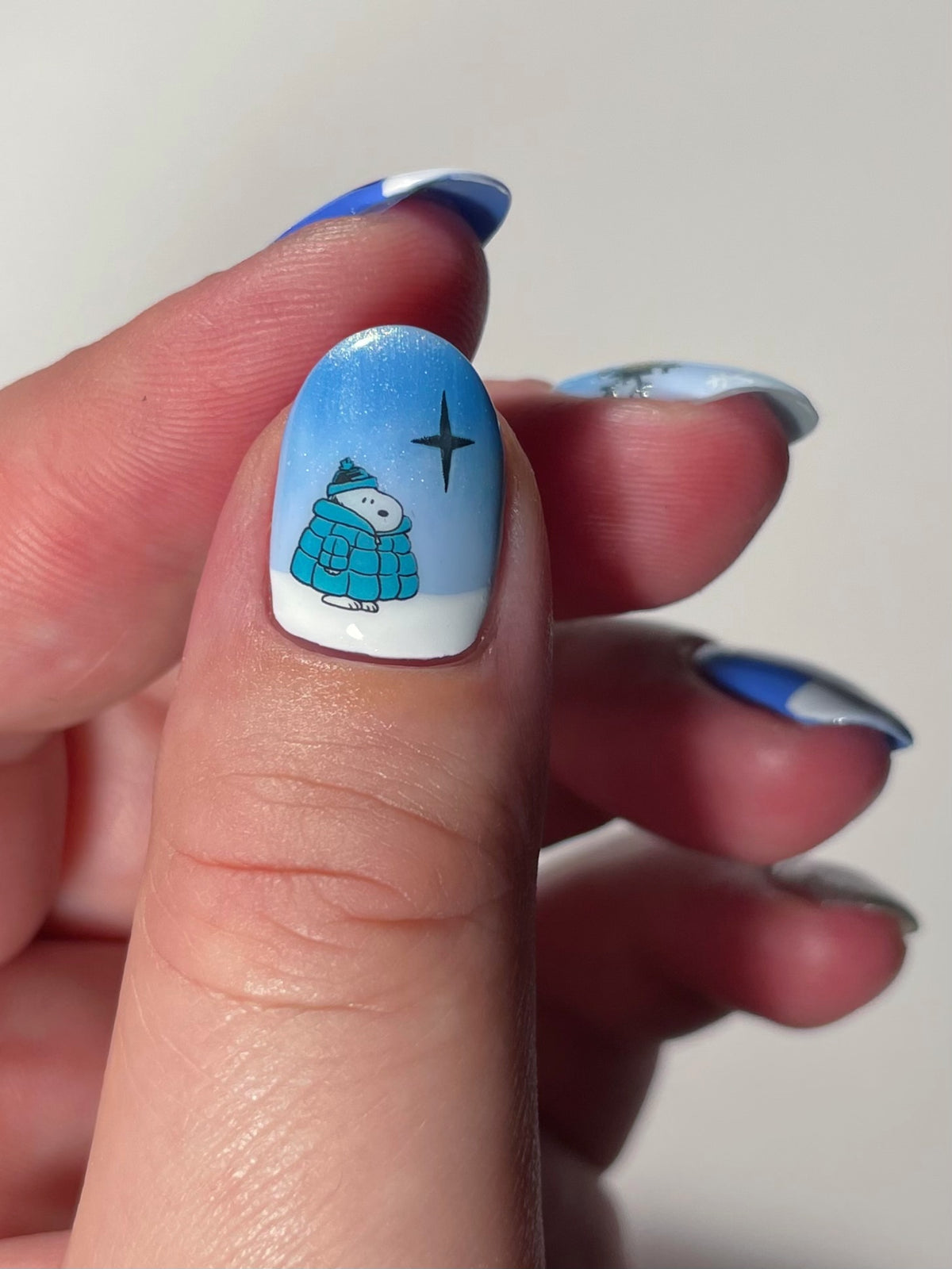 A close-up of a thumb with winter-themed nail art. A light blue ombre nail with nail art stickers of a Snoopy character and silver star.