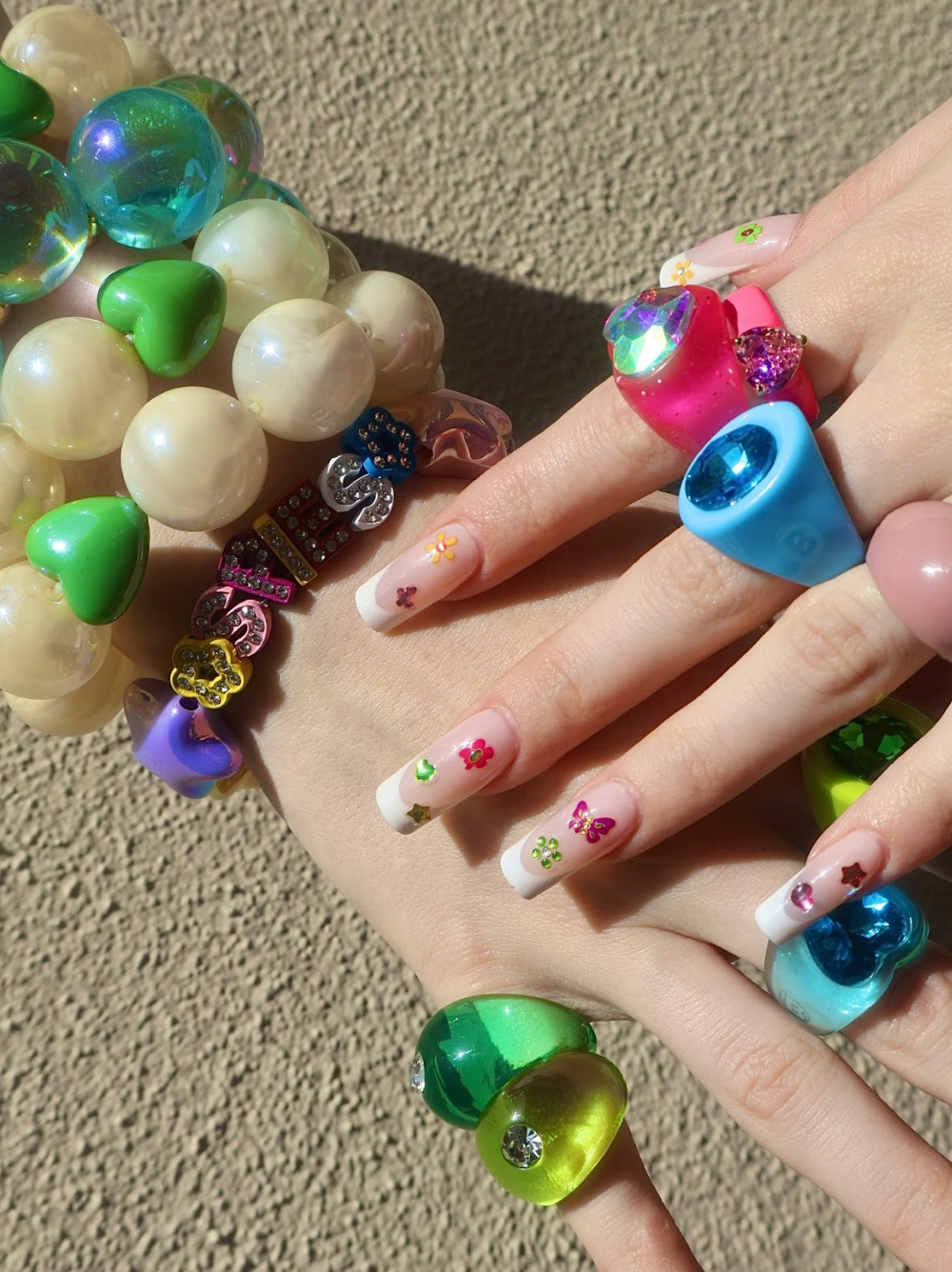 Colorful beaded bracelet on a textured surface with a hand wearing multiple rings and colorful nails art stickers on the nails.