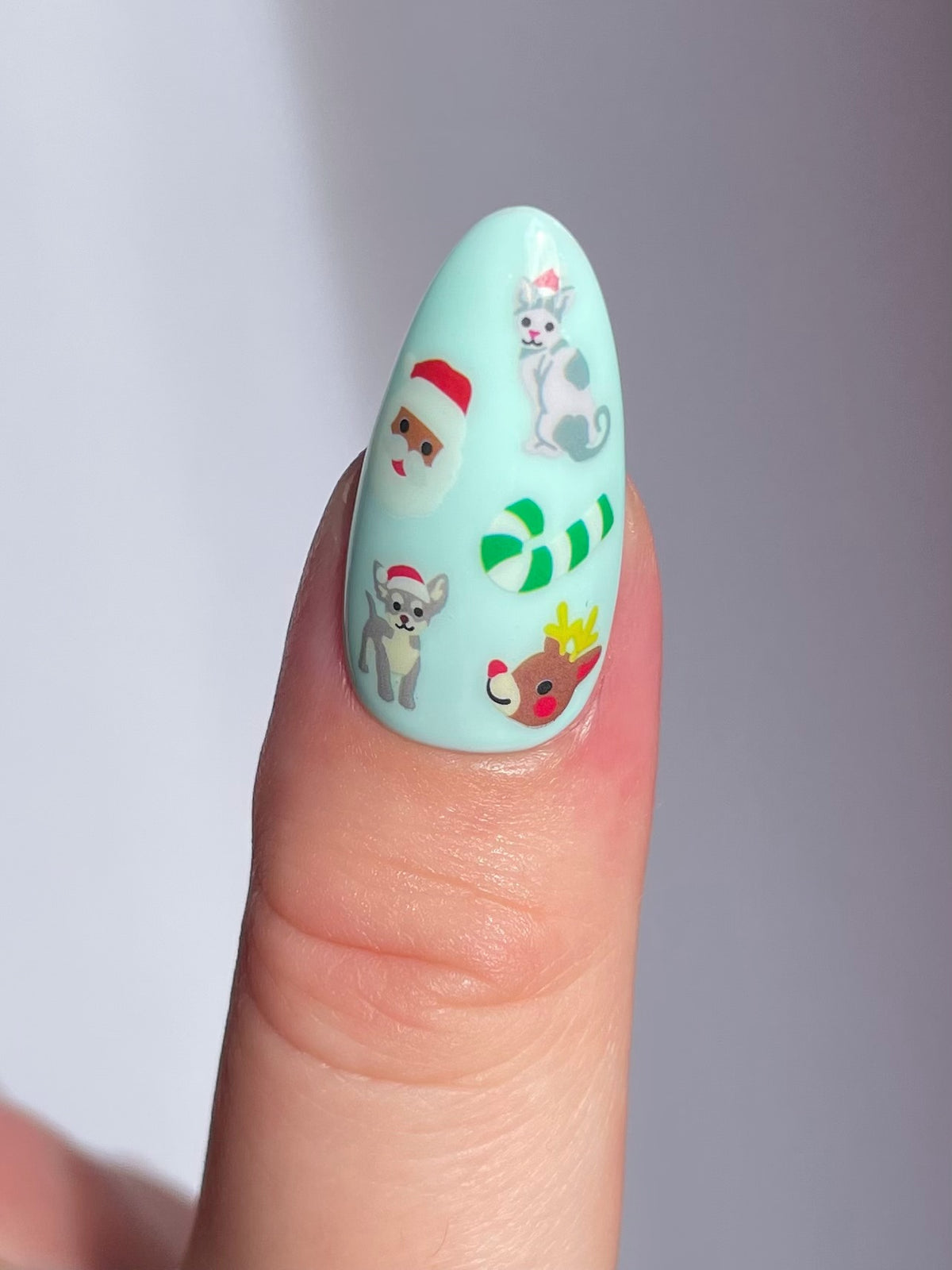 A close-up of a thumb showing long, almond-shaped nail with a light mint green base with holiday-themed nail art stickers.