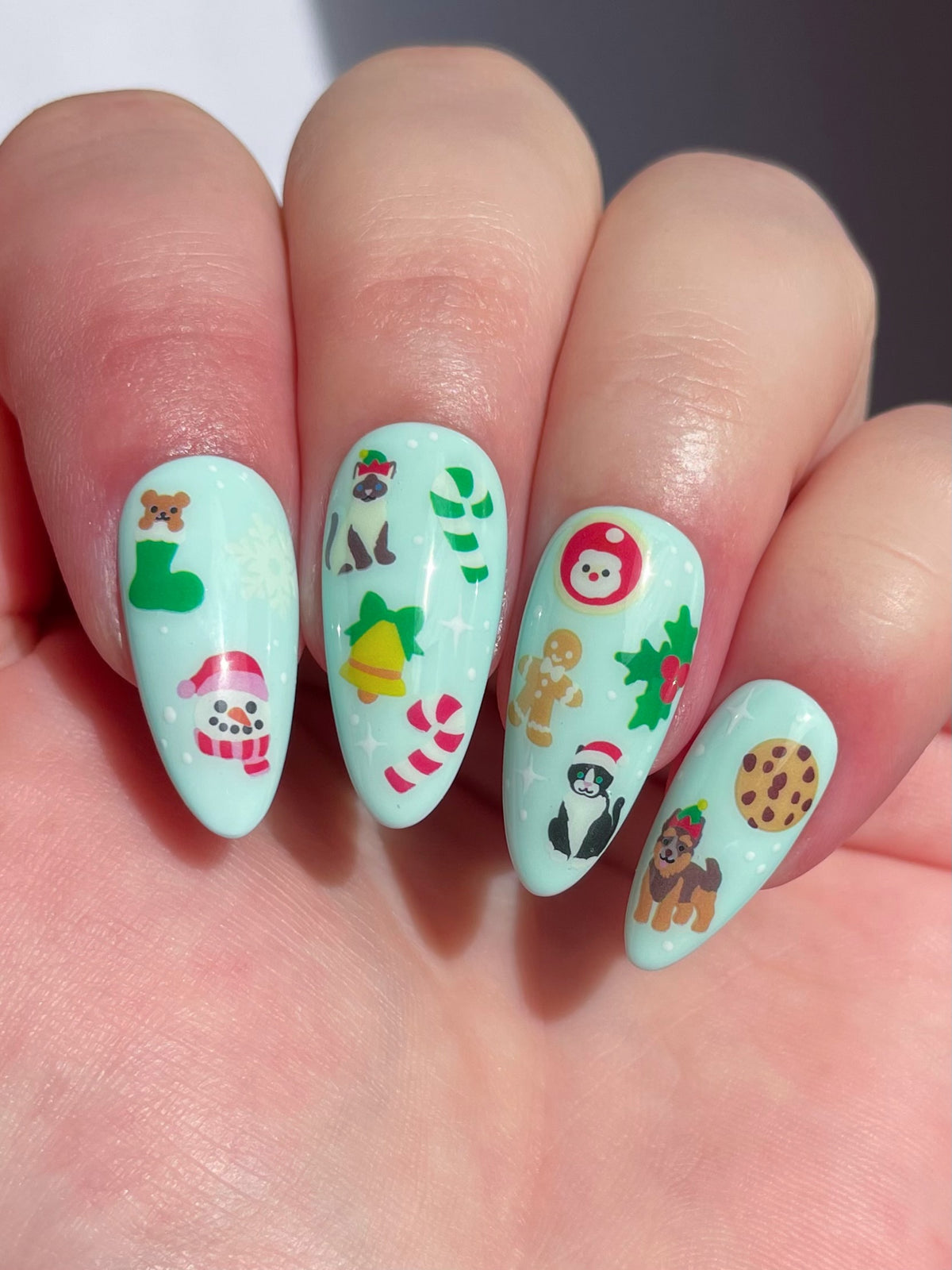 A close-up of a hand showing long, almond-shaped nails with a light mint green base with holiday-themed nail art stickers.