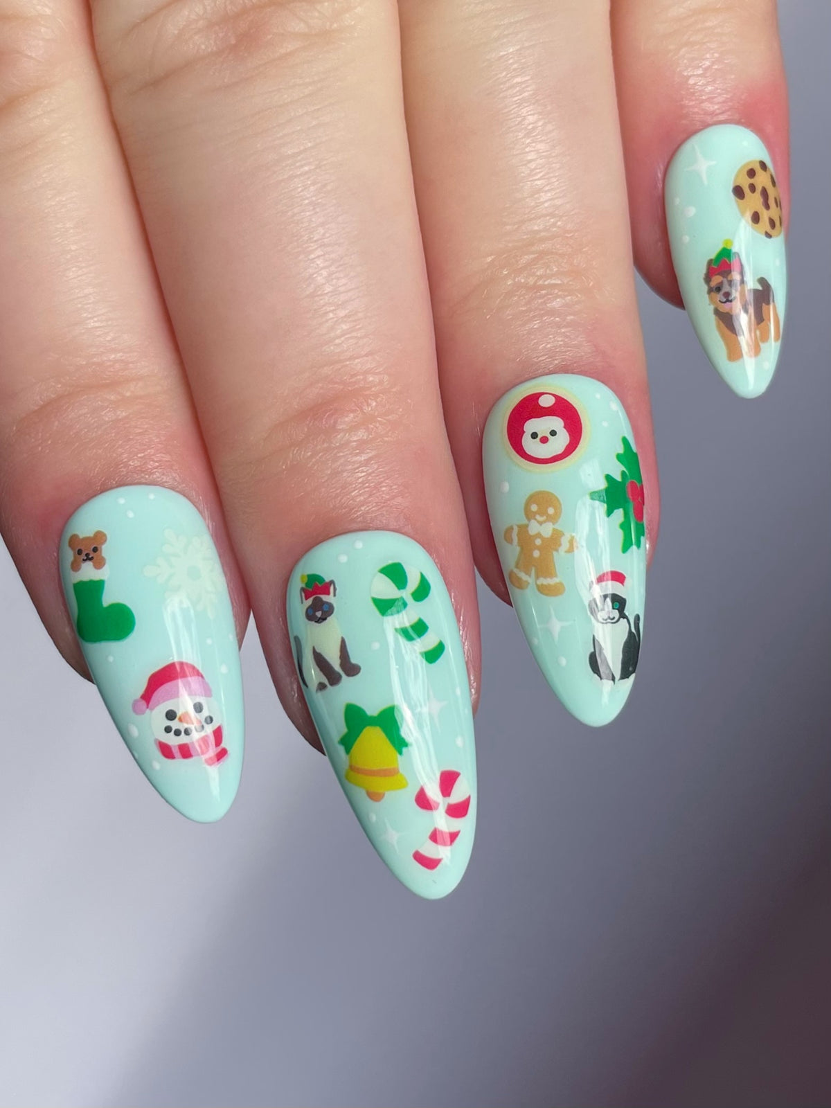 A close-up of a hand showing long, almond-shaped nails with a light mint green base with holiday-themed nail art stickers.