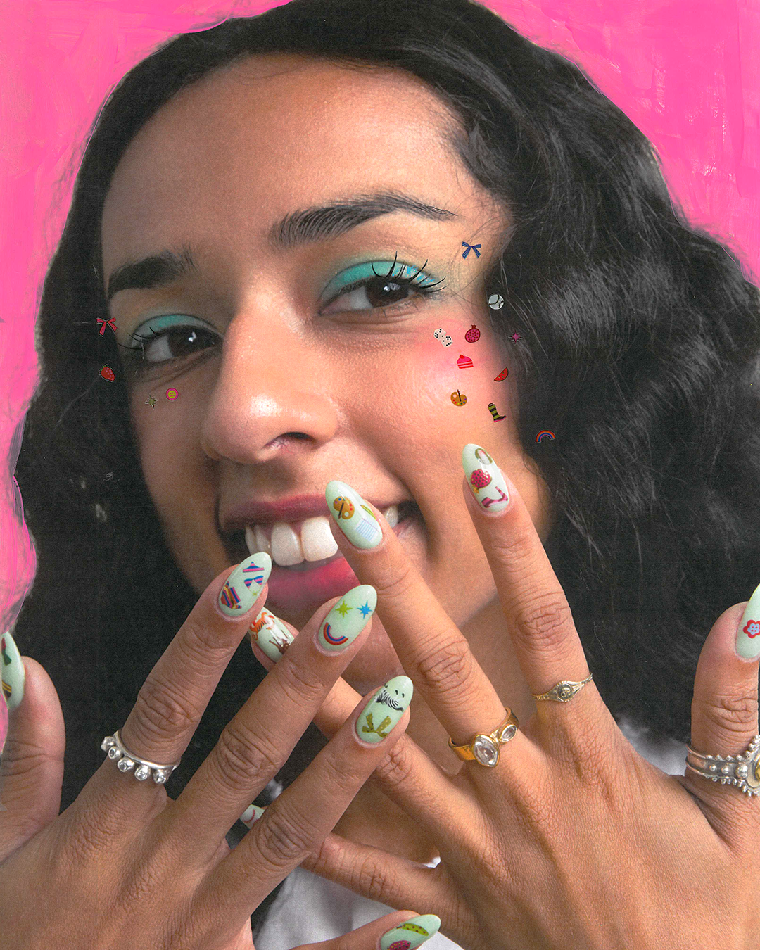 A close-up lifestyle shot of a woman with a bright smile, showing her nails with nail art stickers, along with silver rings, set against a vibrant pink background.