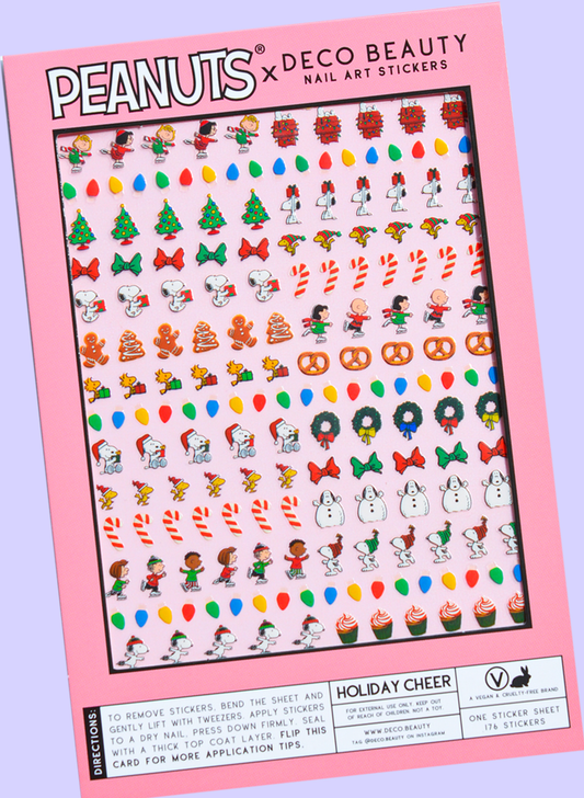 A flat lay of Peanuts x Deco Beauty Holiday Cheer nail art stickers, with festive stickers including Christmas trees, candy canes, snowmen, and Peanuts characters in light pink packaging.