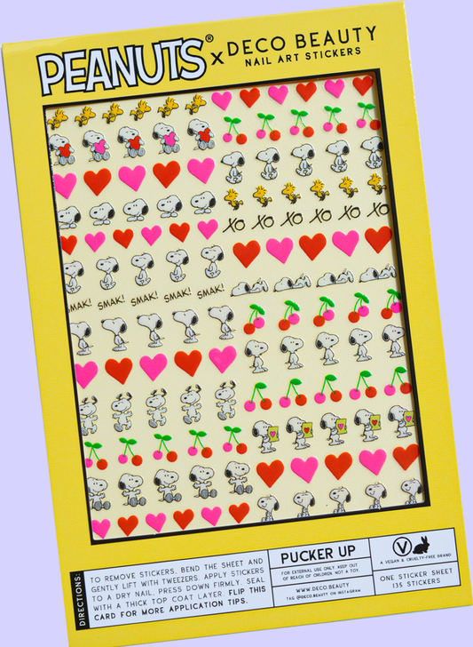 A flat lay of a sheet of Peanuts x Deco Beauty nail art stickers featuring Snoopy, Woodstock, hearts, cherries, and 'xo xo,' in yellow packaging.
