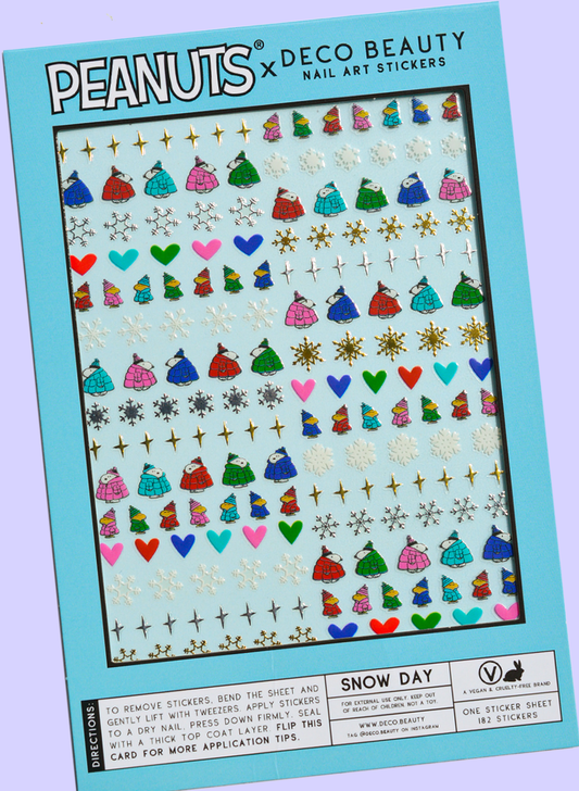 A flat lay of Peanuts x Deco Beauty 'Snow Day' nail art stickers, featuring colorful winter-themed stickers including snowflakes, hearts, Peanuts characters, and stars in light blue packaging.