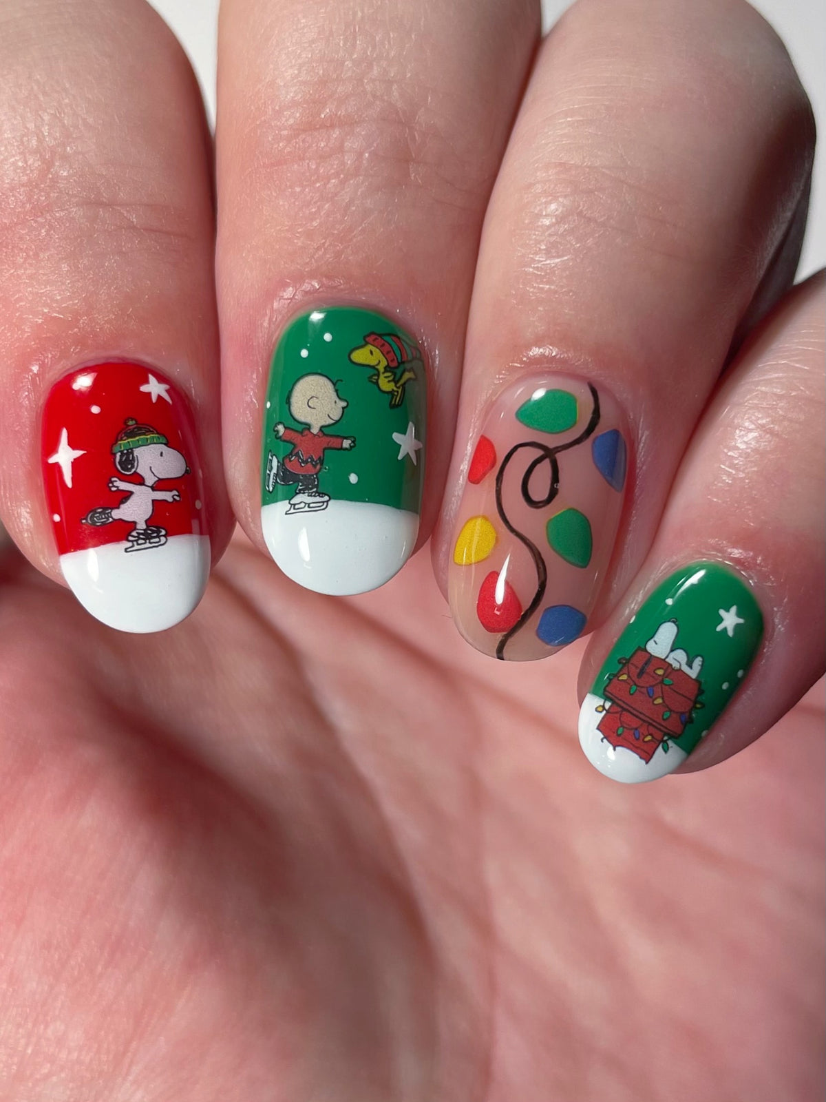 A close-up of a hand with holiday-themed nail art stickers featuring Snoopy and Charlie Brown, a Christmas tree, and colorful lights on a red and green background.