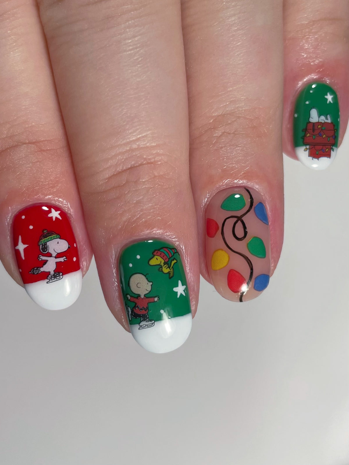 A close-up of a hand with holiday-themed nail art stickers featuring Snoopy and Charlie Brown, a Christmas tree, and colorful lights on a red and green background.