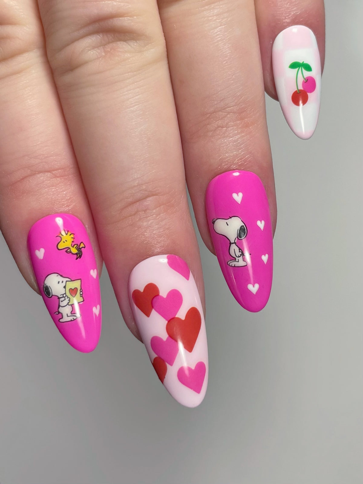 A close-up of a hand with long almond-shaped nails with pink and white nail art stickers, including hearts, Snoopy characters, and cherries.
