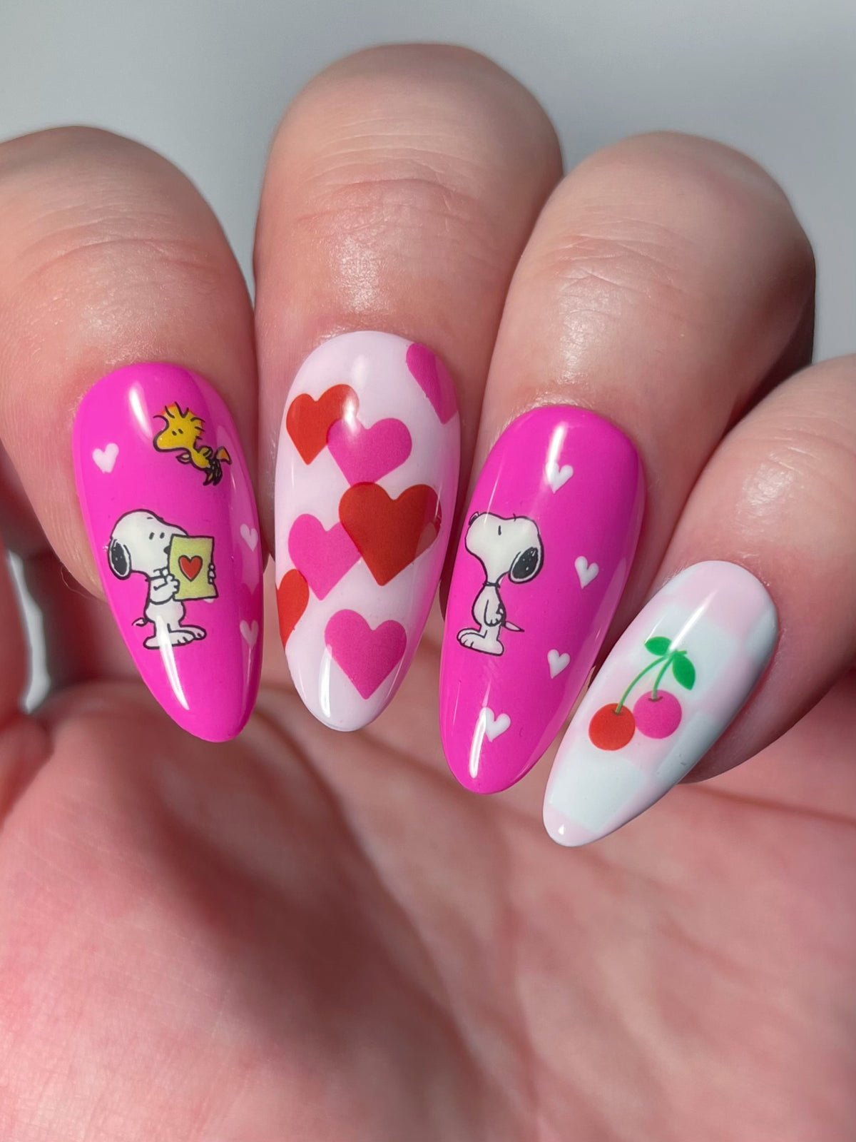 A close-up of a hand with long almond-shaped nails with pink and white nail art stickers, including hearts, Snoopy characters, and cherries.
