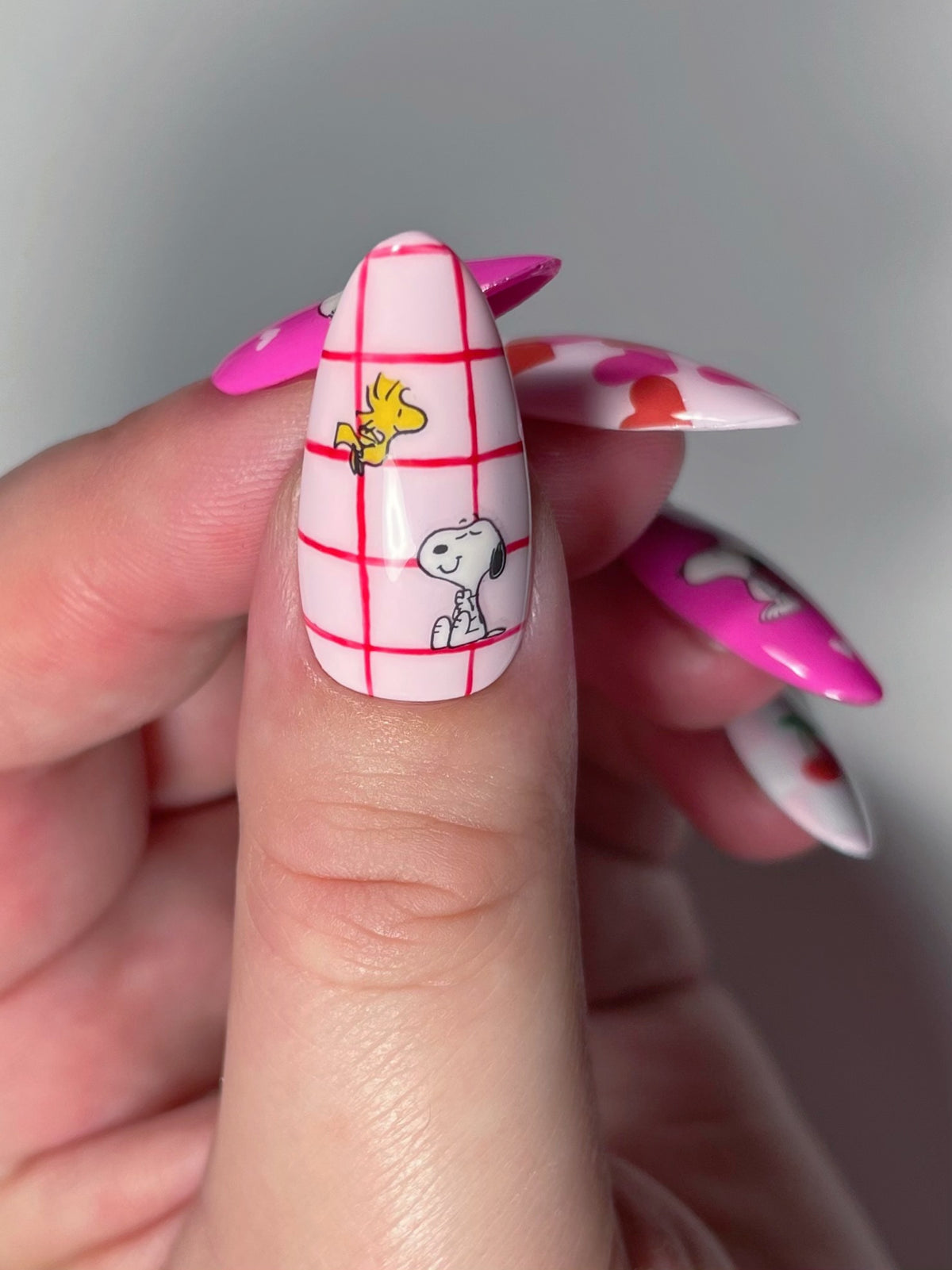 A close-up of an almond shaped thumb nail with a pink and white plaid pattern, with Snoopy and Woodstock nail art stickers.