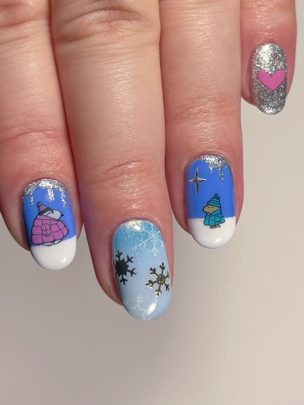 A close-up of a hand with winter-themed nail art. A mix of solid blue, light blue, silver glitter, and white base coats, with nail art stickers of Snoopy characters, snowflakes, and a pink heart.