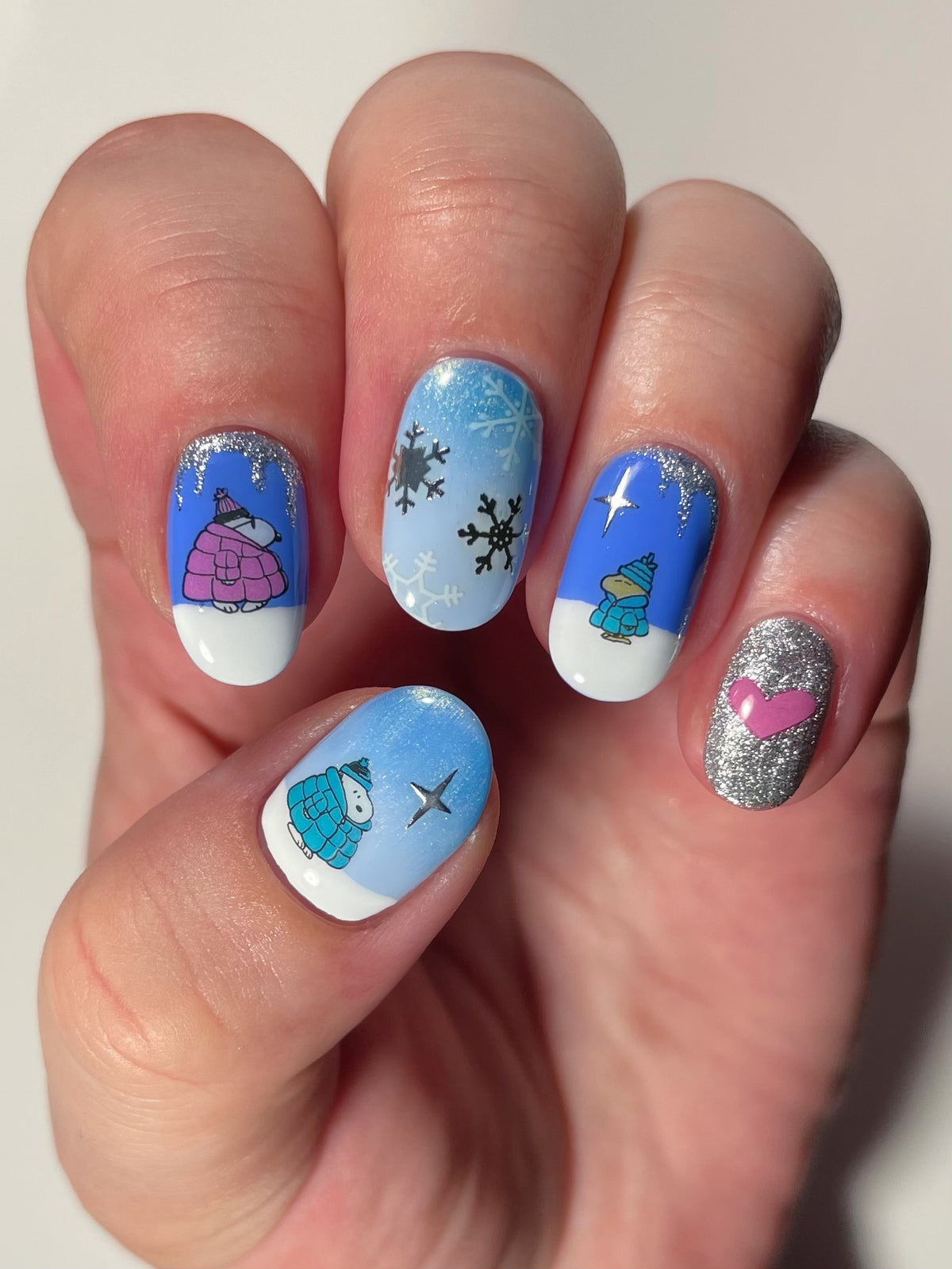 A close-up of a hand with winter-themed nail art. A mix of solid blue, light blue, silver glitter, and white base coats, with nail art stickers of Snoopy characters, snowflakes, and a pink heart.