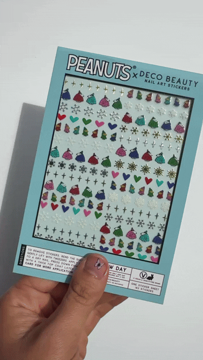 A gif of Peanuts x Deco Beauty 'Snow Day' nail art stickers, featuring colorful winter-themed stickers including snowflakes, hearts, dresses, Peanuts characters, and stars in light blue packaging.