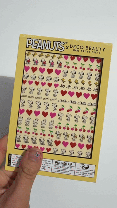 A gif of a sheet of Peanuts x Deco Beauty nail art stickers featuring Snoopy, Woodstock, hearts, cherries, and 'xo xo,' in yellow packaging.